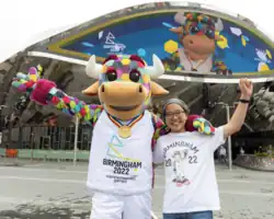 A person wearing a multicoloured bull mascot costume wearing a gold medal and a shirt that says "Birmingham 2022 Commonwealth Games". The person has their left arm around the shoulder of a young girl with glasses. The two are standing in front of New Street Station with a large digital screen displaying an image of the mascot.