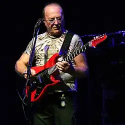 Petr Janda wearing dark pants and a tiger-print t-shirt, playing a red electric guitar on a darkened stage