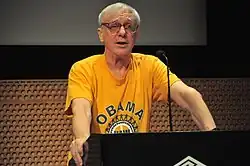 A gray-haired man with glasses wearing a yellow shirt standing at a podium