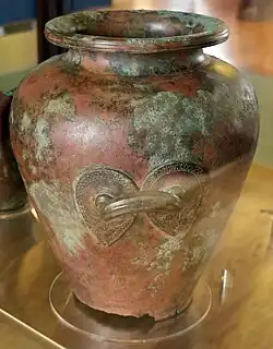Metal vessel with heavy patina: we see it from the side, focusing on a handle whose two ends have been decorated with sculptures of shields.