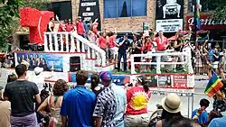 Multiple people on a parade float passing a street lined with people. At the back of the float, left of the image, is a large red T representing the network logo. Several banners promoting Telemundo Chicago programming and diversity initiatives are attached to the skirt of the float.