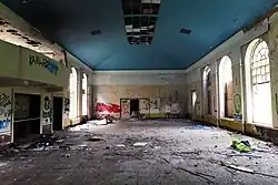 Dance hall like room with high ceiling and high arched windows in dilapidated condition with graffiti, debris and litter