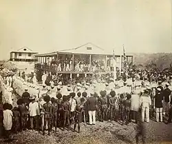 Sepia photo of a large crowd around a two-story building, with two raised flags in front of the building