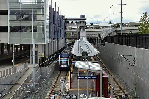 Redmond Technology station on opening weekend in April 2024. The station has an island platform between two tracks. There is a train at the platform. The platform is covered and is located next to a large parking garage. The train is white with blue accents and has a curved front.