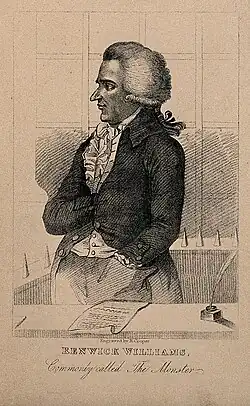 Three-quarter portrait of Williams in the dock. He wears a cropped jacket and has his hand tucked into the breast.