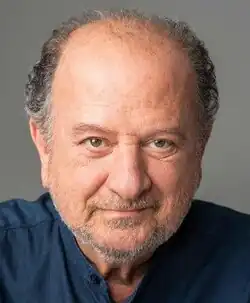 Colour publicity headshot of a half-smiling man with short greying hair brushed back, designer stubble and brown or hazel eyes