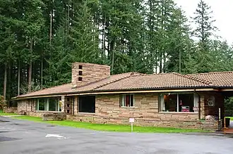 Brown one-story modern flagstone building, parking lot in front and fir trees behind it.