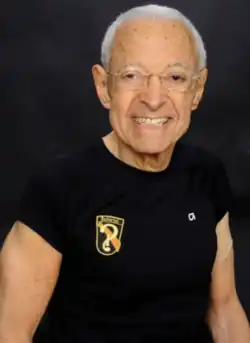 Caucasian male with shortwhite hair and wire-rimmed glasses, smiling; wearing black short-sleeved tee-shirt