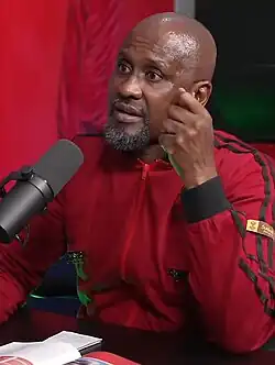 A man seated at a table and behind a microphone, wearing a red tracksuit with black stripes