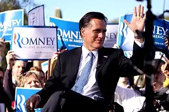 Mitt Romney sitting outdoors during daytime, with crowd behind him holding up blue and white "Romney" signs
