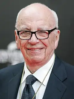 An old man with a bald head and a black suit, as well as glasses, is standing, smiling.