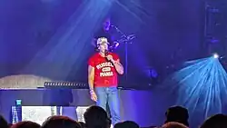 A caucasian man on stage in a red shirt and blue jeans