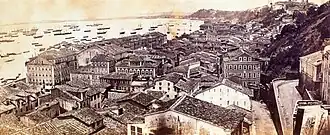 Photograph looking down a hillside overlooking the buildings of a town with a harbor beyond which is crowded with various vessels