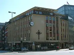 A 1950s or 1960s six-story building, predominantly made of yellow brick with red brick used around the windows. Other ornamentation include a large round black and white clock, a large black cross symbol, and "Scientology Kirche Hamburg e.V." lettering.