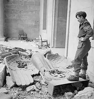 Achromatic photograph of a soldier standing next to a fallen Nazi eagle statue, on which a dark linen rests, in a ruined courtyard