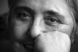 A close-up black-and-white portrait of an older woman smiling at the camera. Her fist is resting against her cheek, obscuring half her mouth.