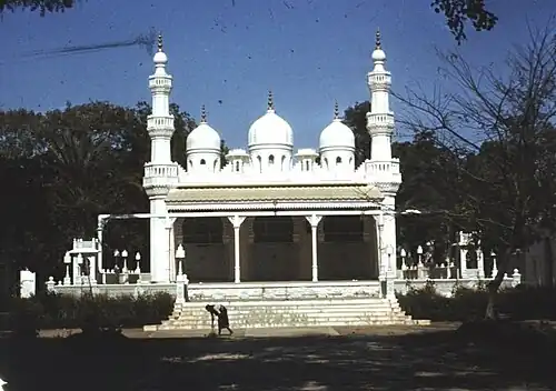 A white mosque with three domes, flanked by two minarets on the sides. Trees are seen in the background.