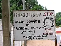 A fading sign that says: GAMCOTRAP - Gambia Committee on Traditional Practices. 49 Garba Jahumpa Road, Bakau New Town. There is a phone next to the numbers 497416. On the right side of the sign, there is a drawing of a crying woman. Above her, it says STOP; below, it says FEMALE GENITAL MUTILATION. This text is in red.