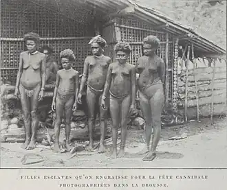 Black-and-white photograph of five nearly naked young women and girls standing in front of a hut; a sixth is visible in the background.