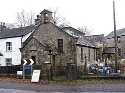 A small stone church with a porch and a bellcote in the foreground, and the body of the church, on which is a glazed lantern on the roof, receding into the background