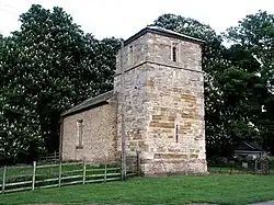 A simple stone church showing a stubby tower with pyramidal roof and lancet windows and beyond that, the nave with a single window