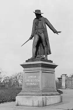 A statue of a man wearing cape and hat, and holding a sword standing to the left