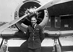 Black and white photograph of a man in an air force uniform, standing in front of the propeller of an aircraft, near its front landing wheels.