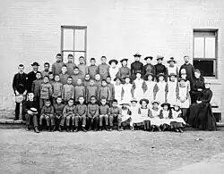 Posed, group photo of students and teachers, dressed in black and white, outside Middlechurch, Manitoba's St. Paul's Indian Industrial School