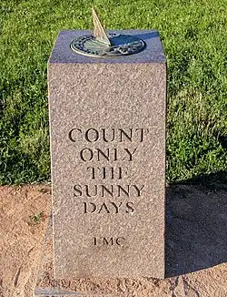 A sundial rests on a marble rectangular prism which is inscribed with the words, "Count only the sunny days." It is golden hour.