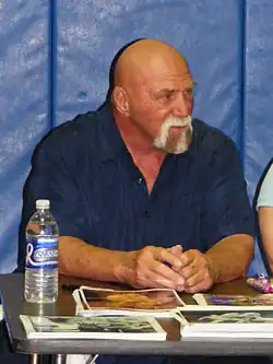 Balding sixty-year-old man wearing a partially-grey goatee and dark blue shirt. He is looking towards the right and sitting at a table with stacks of photos in front of him and a bottle of water to the left.