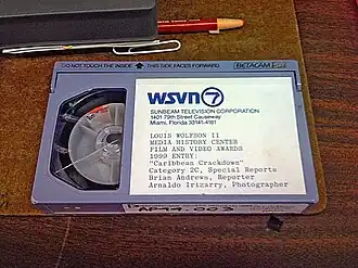 A videocassette on top of a clipboard next to its plastic case, a grease pencil and a paper clip. The label on the cassette contains the WSVN letterhead: "WSVN 7 Sunbeam Television Corporation, 1401 79th Street Causeway, Miami, Florida 33141-4181" then in print below, "LOUIS WOLFSON II MEDIA HISTORY CENTER FILM AND VIDEO AWARDS 1989 ENTRY: 'Caribbean Crackdown' Category 2C, Special Reports, Brian Andrews, Reporter, Arnaldo Irizarry, Photgrapher"