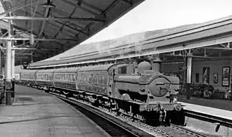A pannier tank locomotive pulling five passenger coaches through a station