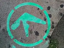 The edge of the pavement on a city street in New York, United States. Green-coloured spray-painted graffiti depicts the Aphex Twin logo inside a dashed outer circle. At the bottom right is roadside greenery.