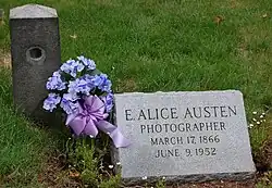 A headstone reading "E. Alice Austen, Photographer, March 17, 1866, June 9, 1952. There are blue and purple flowers with a purple bow beside it.