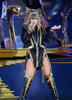 Taylor Swift singing on a microphone, dressed in a black suit with golden accents