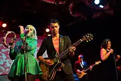 A band performing on a stage. From left to right; singer and percussionist, guitarist, bass guitarist and another singer.