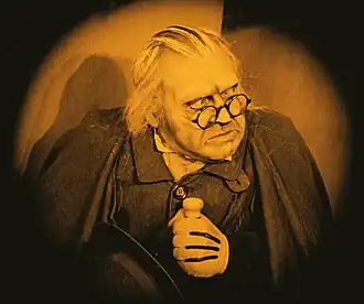 A man wearing round-framed glasses, wearing a black coat and shirt, and white gloves with black lines along the knuckle lines. The man is looking off camera with a concerned look on his face.