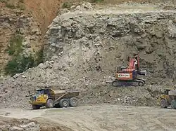 A photograph showing quarrying operations with a mechanical digger in the centre extracting rock from a cliff face. To the left, a lorry is just leaving with a full load of rock, and to the right, another lorry is waiting to collect its load from the digger.