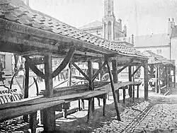 Monochrome photograph of the cobblestone alley behind the shambles. The shambles have pantile roofs, benches, and curved roof trusses. The market cross can be seen in the background.