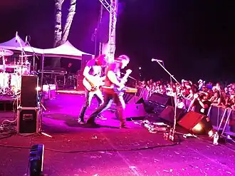 Side of stage shot of a band playing. Two members are shown in right profile both are playing guitars. Other stage equipment is visible around them. The audience is to the right and below stage level.