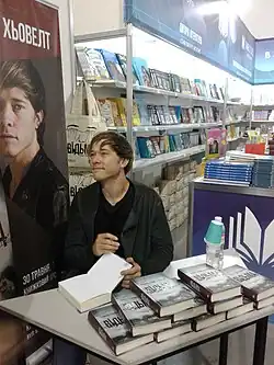 Thomas Olde Heuvelt wearing a black t-shirt and dark jacket, signing a book at a table in a bookstore, looking up and smiling, looking right of camera