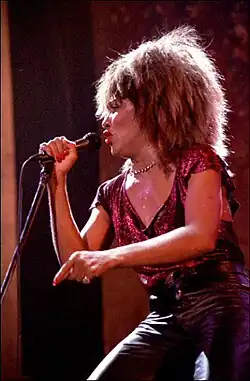 Tina Turner with a red top and black jeans in her mid-forties, singing into a microphone on stage
