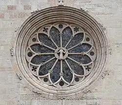 Photograph of a grey stone wall. There is a circular window. In the middle, a standing figure in a robe raises its arms to the sides to reach a circle. From the circle, twelve columns radiate. The ends of the columns are joined by arches. Outside of the frame of the window, twelve figures of people. The figure on top sits on a throne but is not totally visible in this photograph.