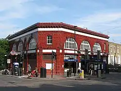 A two-storey station building in red glazed blocks sits at the corner of a road junction. Large semi-circular windows feature prominently on the upper floor.