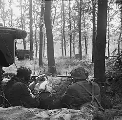 Two men armed with Sten guns, facing away from the camera in woods