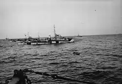 An inflatable raft on the water with four camouflaged World War II-era warships behind it. No land is visible.