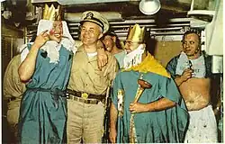 Color photograph of crossing the line ceremony held in the crews mess compartment of the nuclear submarine USS Triton that includes (from left to right) crewman Wilmot A. Jones dressed as the Queen of the Royal Court; Captain Edward L. Beach dressed in khaki uniform and ceremonial sword; Chief Loyd Garlock dressed as King Neptune, Ruler of the Raging Main; crewman Ross MacGregor dressed as Davy Jones in background; and crewman Harry Olsen dressed as the Royal Baby.