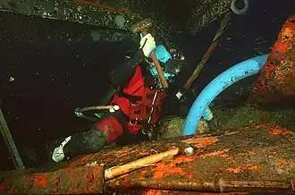 A diver wearing lightweight helmet and hot-water suit working on a piece of corroded and encrusted wreckage with a club hammer