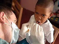 Man holding plastic tray with brown material in it and sticking a small stick into a boy's open mouth