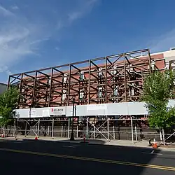 A partially constructed building with exposed steel framework and brick walls, surrounded by scaffolding. Trees are visible in front, and the scene shows a clear blue sky above.